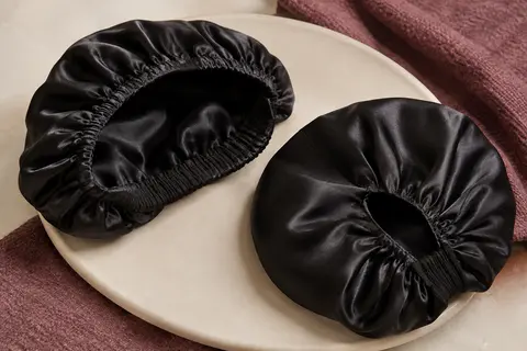 Deux bonnets de nuit noirs en satin posés sur une céramique mate, avec leur bande élastique visible.