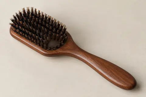 Brosse plate en bois brun avec manche allongé et tête garnie de poils, posée sur une céramique mate.