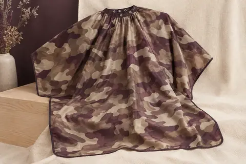 Cape de barbier en polyester à motif camouflage, étendue sur un support en bois clair dans une lumière douce.