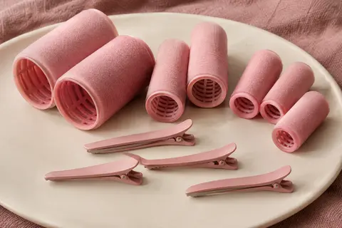 Bigoudis roses sans chaleur en trois diamètres avec des pinces, disposés sur un support en céramique mate.