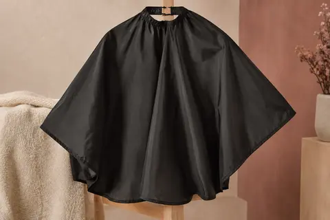 Cape de coiffure noire en nylon, étalée sur un support en bois clair avec sa fermeture à crochets visible