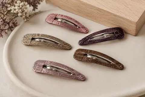 Barrettes à pression en métal pailleté, aux coloris assortis, disposées sur un support en céramique mate.