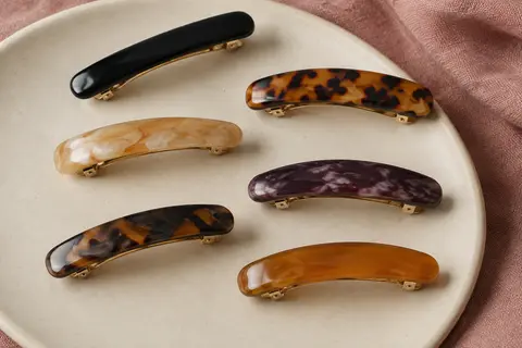 Six barrettes automatiques en acétate aux tons variés, disposées sur un support en céramique mate.