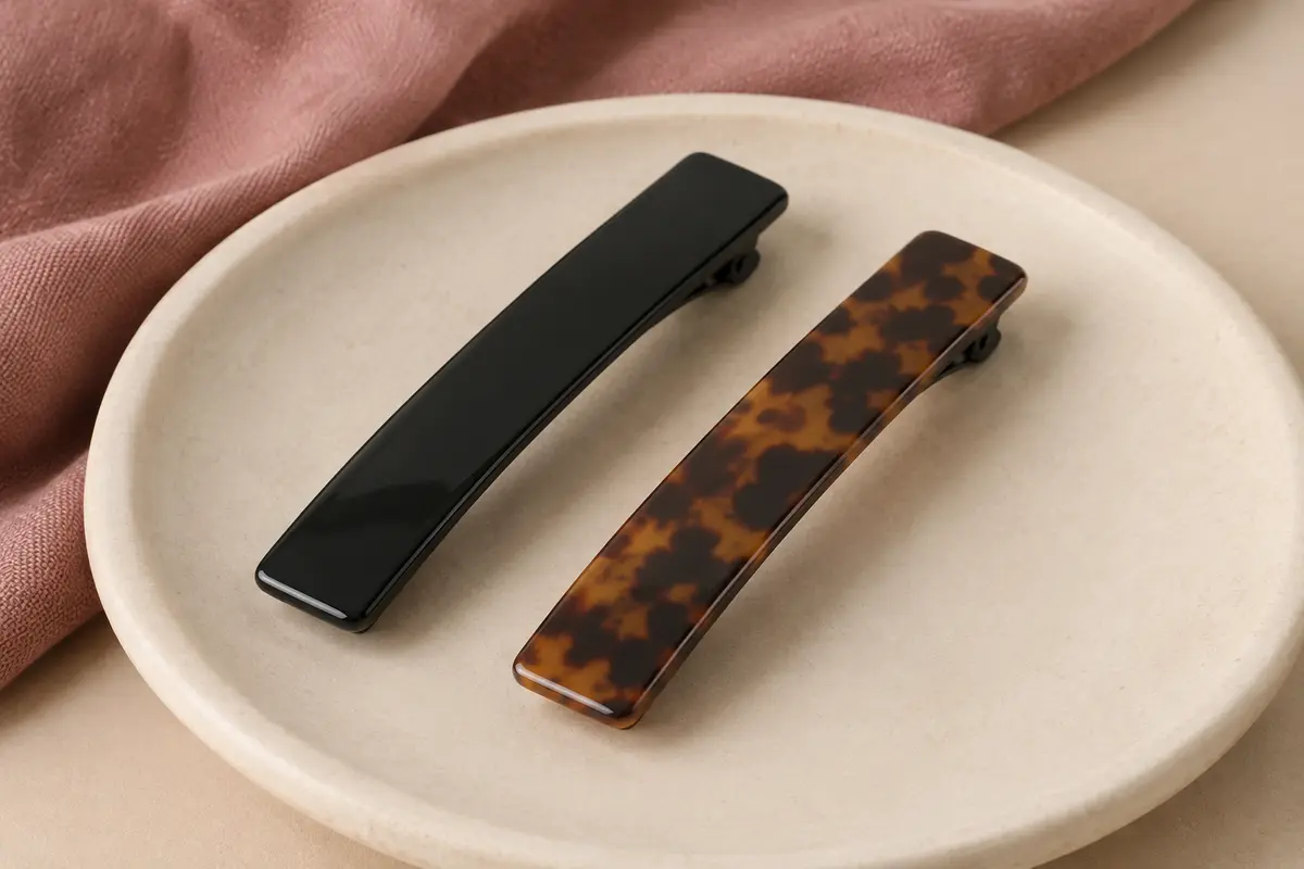 Deux pinces à cheveux plates en plastique, l’une noire et l’autre à motif écaille, posées sur de la céramique mate.