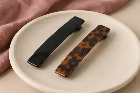 Deux pinces à cheveux plates en plastique, l’une noire et l’autre à motif écaille, posées sur de la céramique mate.