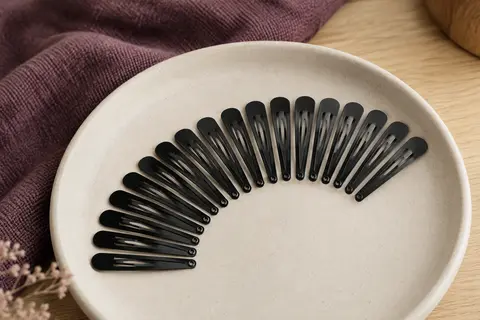 Vingt barrettes plates en métal noir disposées en éventail sur un support en céramique mate crème.