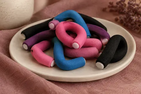 Bigoudis souples en mousse bleus, roses et noirs disposés sur un support crème et un tissu texturé.