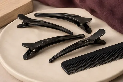 Pinces de séparation en métal noir à bec de canard, accompagnées d’un peigne de coiffage sur céramique mate.