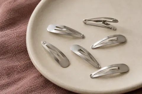 Lot de barrettes à pression en métal argenté disposées sur une céramique mate et un tissu vieux rose.