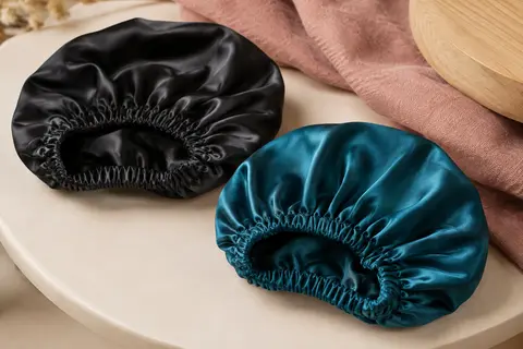 Deux bonnets de nuit en satin de soie noir et bleu paon posés sur un support en céramique mate.