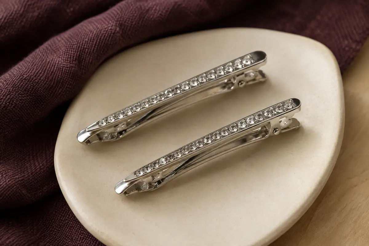 Deux barrettes métalliques argentées ornées de petits strass, posées sur un support en céramique mate.